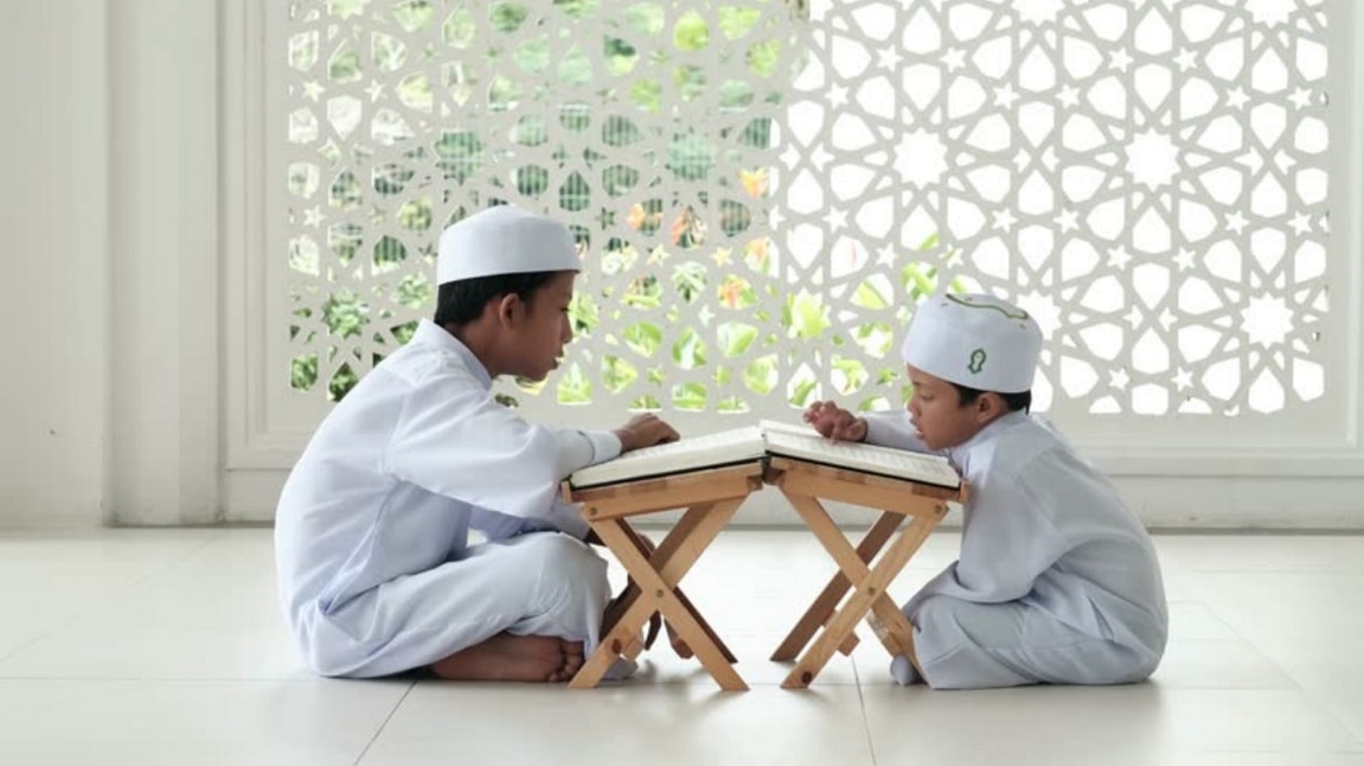 Two young children, one younger than the other, wearing white clothes and hats, sitting cross-legged and facing one another. They each have a small table in front of them with a large book on top. Both children are reading from the books.