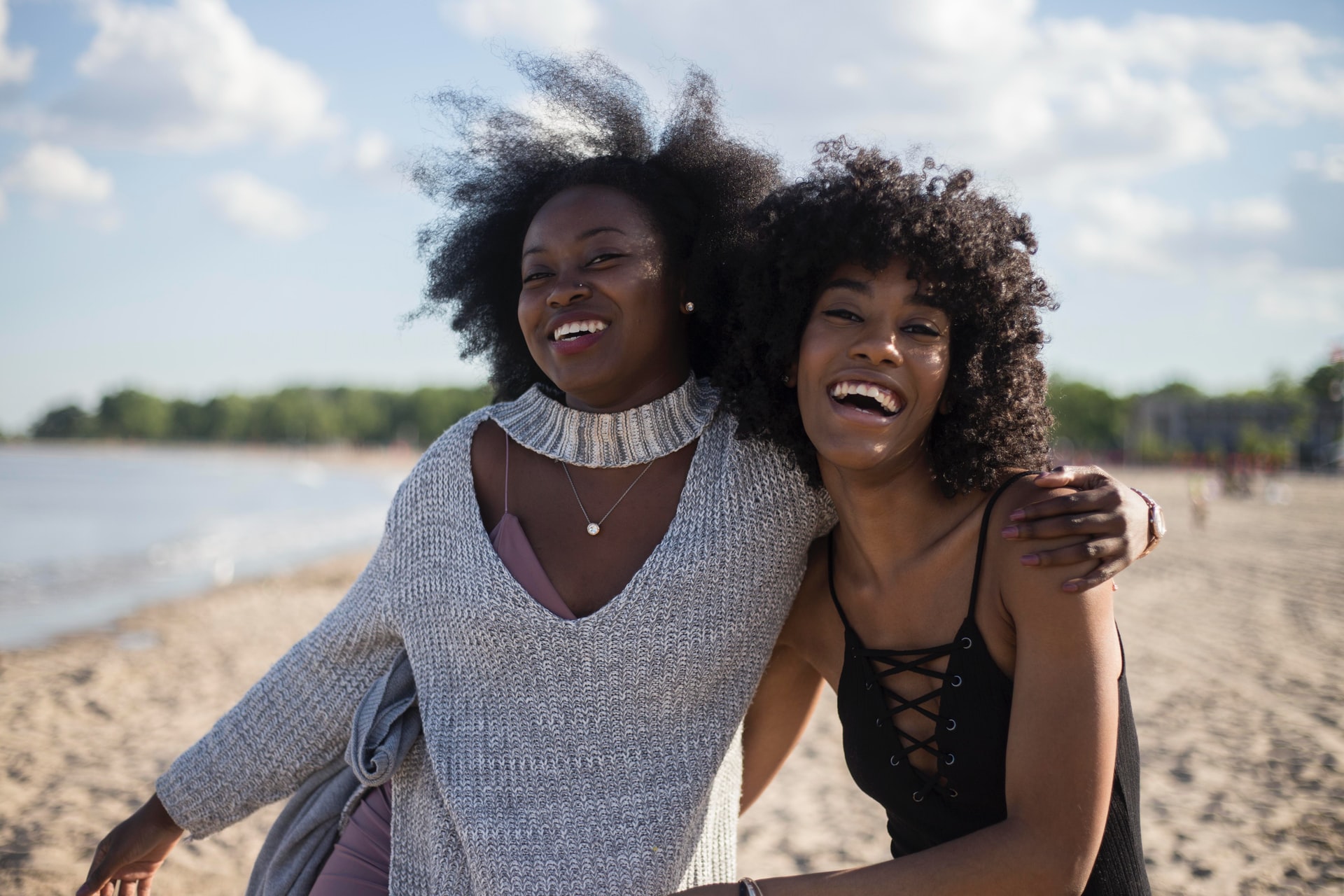 Two people with dark brown skin and dark, curly hair, smiling on a beach. Each person has their arm around the other.